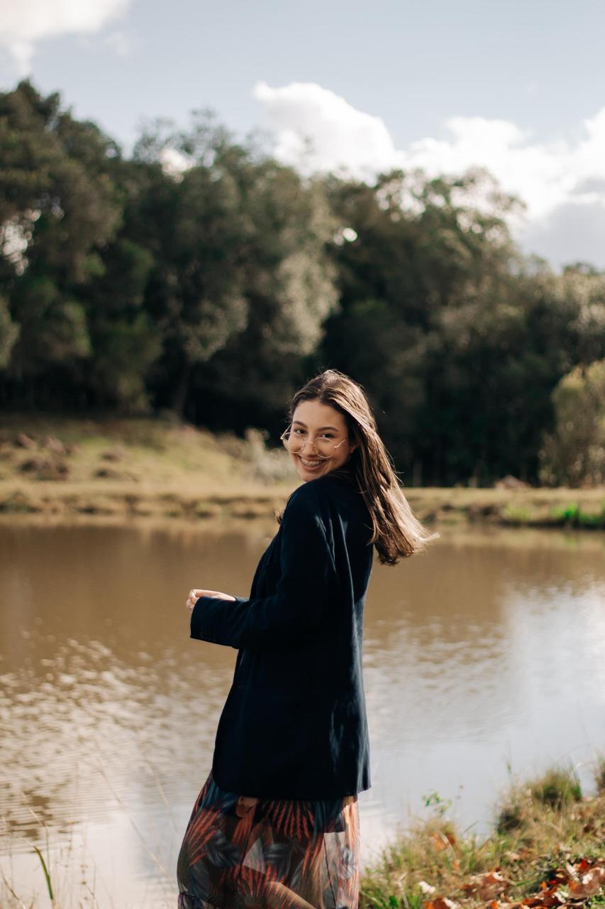 Mylenna sorrindo à beira de um lago, cercada pela natureza